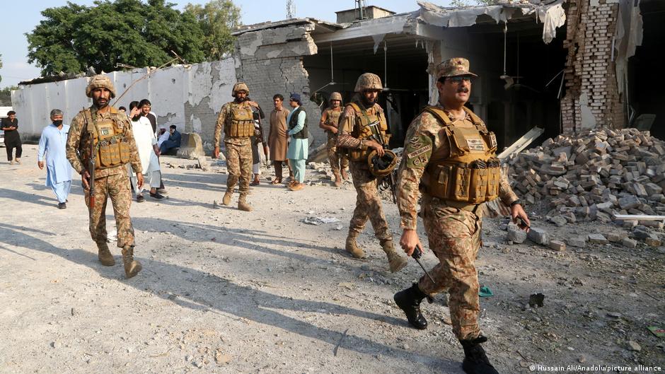 Uniformed armed personnel walk past a damaged building and scattered rubble as civilians stand nearby.