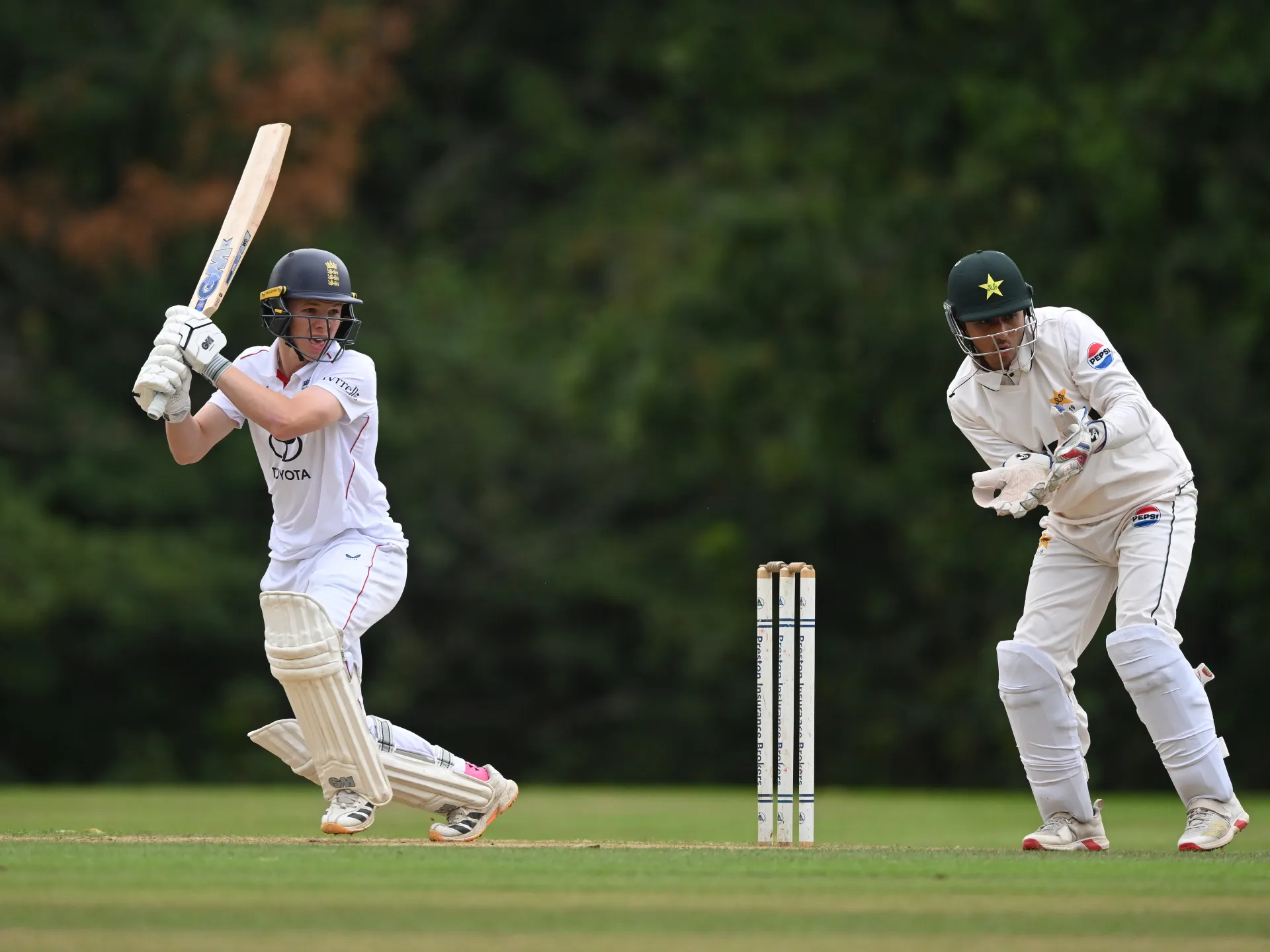 Two cricket batsmen in white uniforms play on a grassy pitch, with stumps between them.