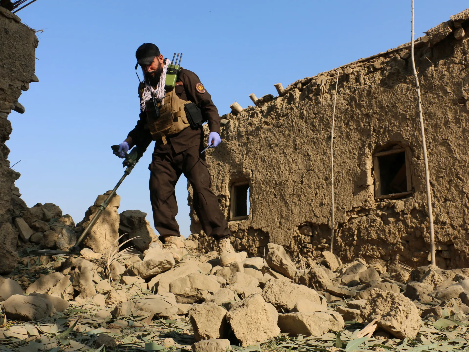 Armed man in tactical clothing stands amid rubble beside a damaged stone building.