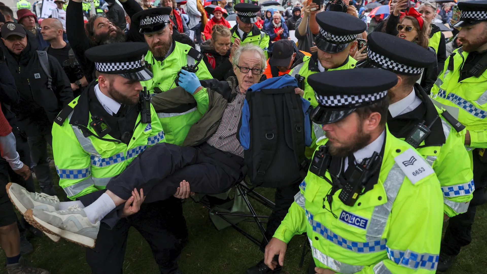 Police officers carry a man through a dense crowd of protesters, with bystanders watching and recording.