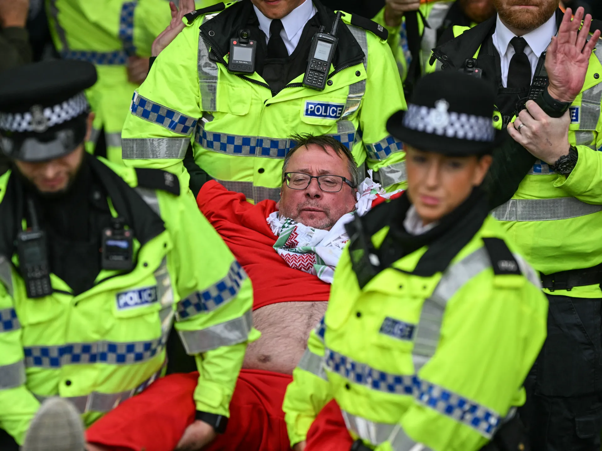 Police officers carry a man in a red top and patterned scarf through a crowd.