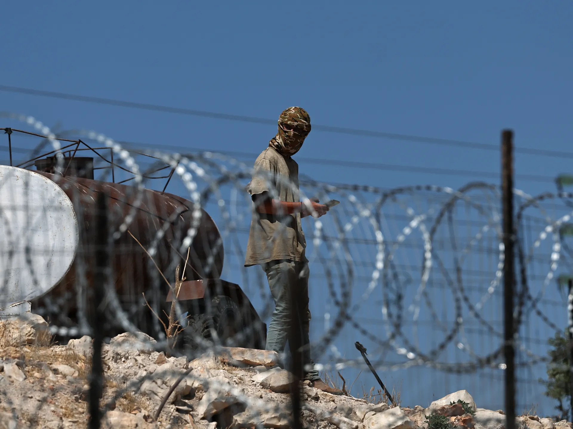 Masked person stands behind coils of barbed wire near rusted machinery on rocky ground.