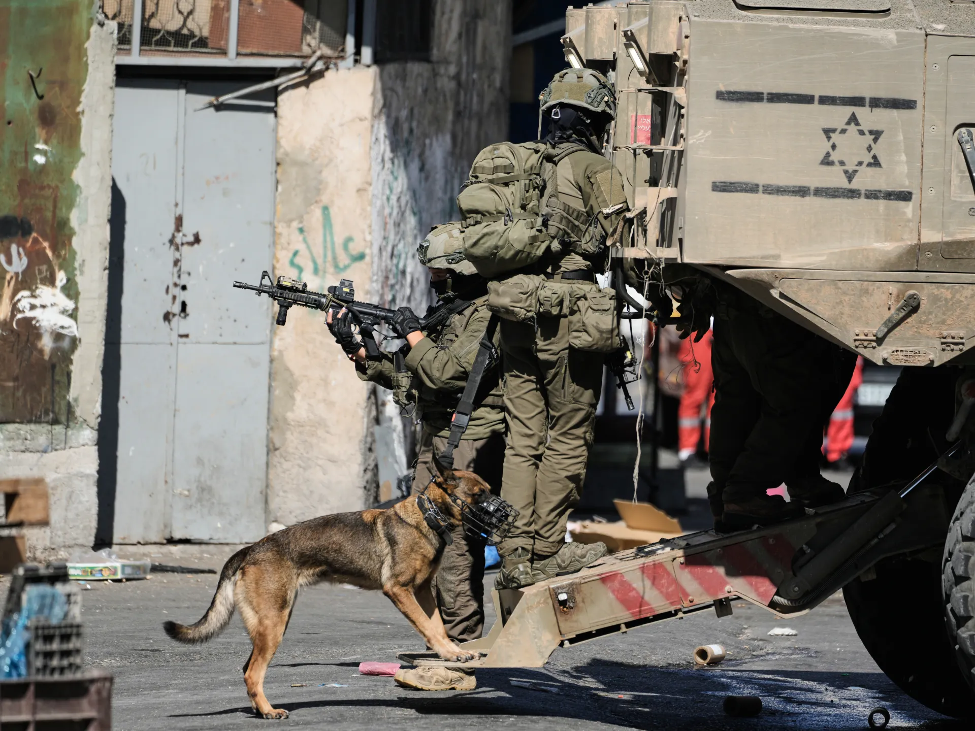 Armed soldiers and a muzzled military dog stand beside an armored vehicle on an urban street.