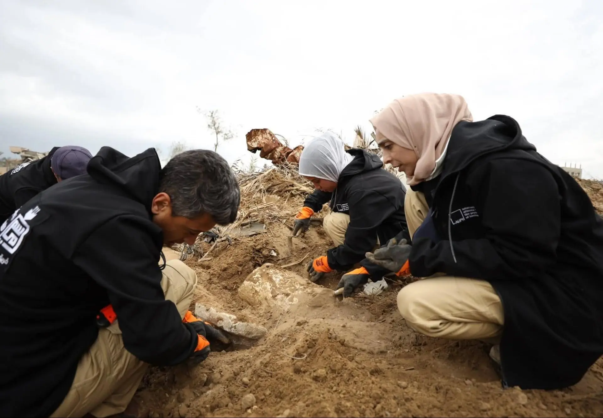 Archaeologists in gloves kneel and carefully uncover stones amid sandy rubble beneath a cloudy sky.