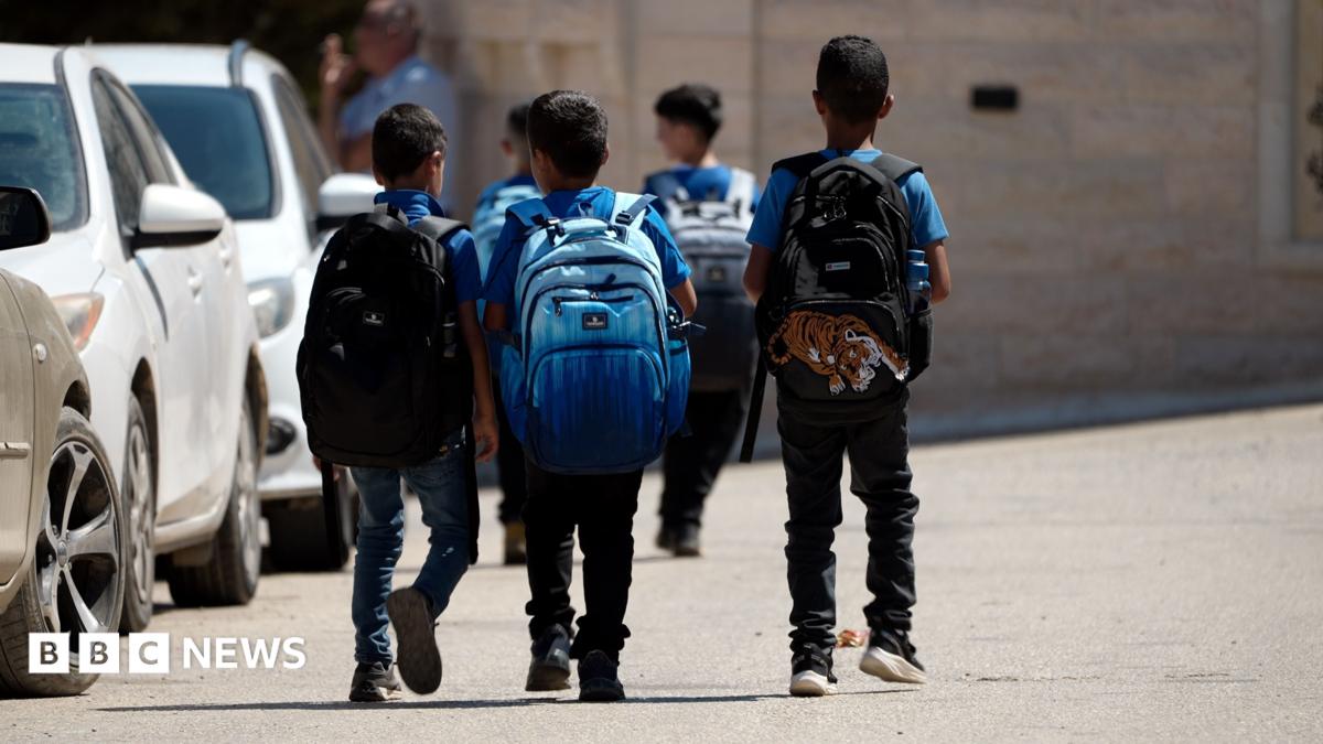 Three schoolchildren with backpacks walk along a street past parked cars.