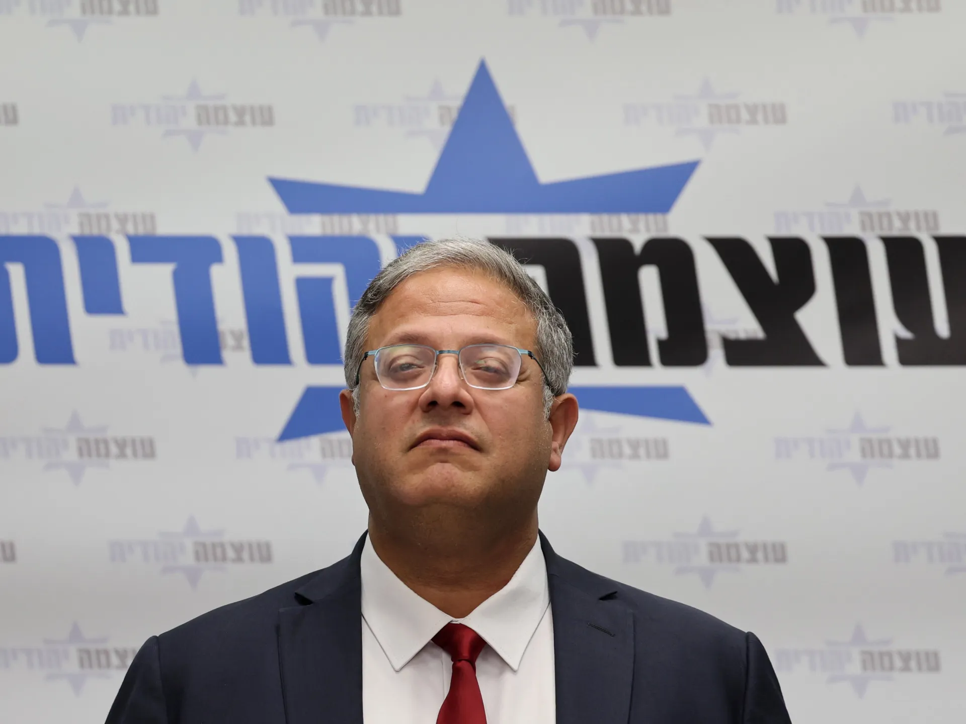 A bespectacled man in a dark suit and red tie stands before a blue-and-black Hebrew party backdrop.