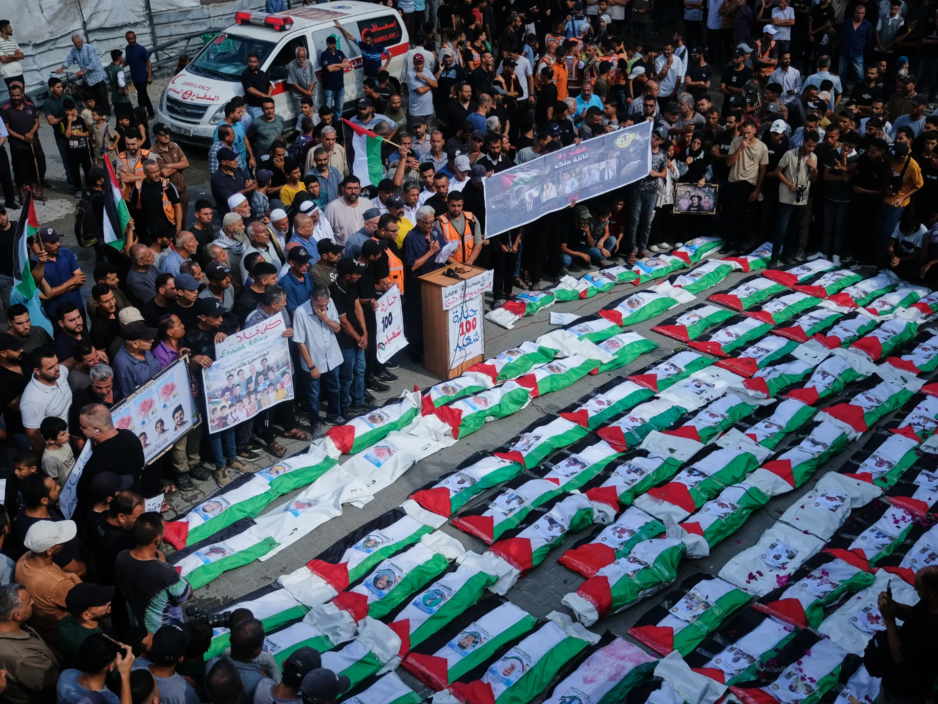 Crowd gathers beside rows of Palestinian-flag-draped bodies during a funeral procession.