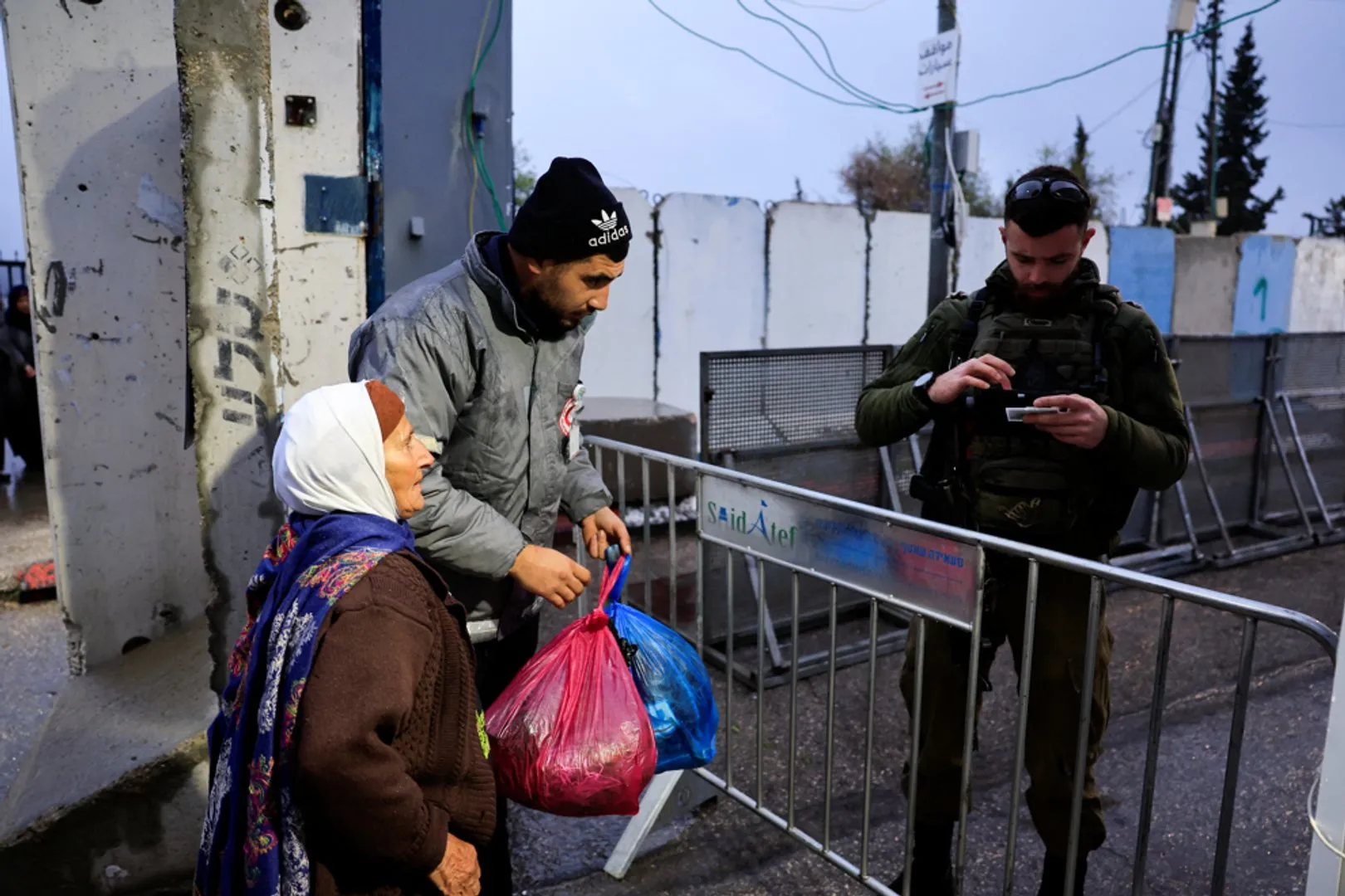 Palestinian woman and man carrying bags stand near metal barriers beside an armed man using a phone.