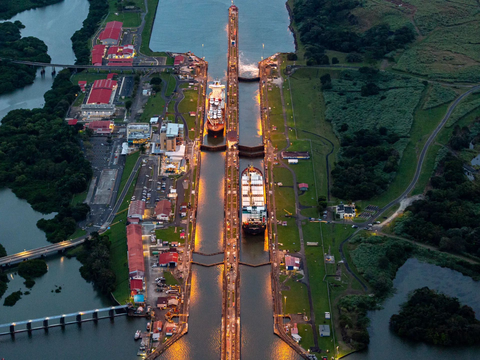 Aerial view of canal locks with two cargo ships passing through amid surrounding waterways and greenery.