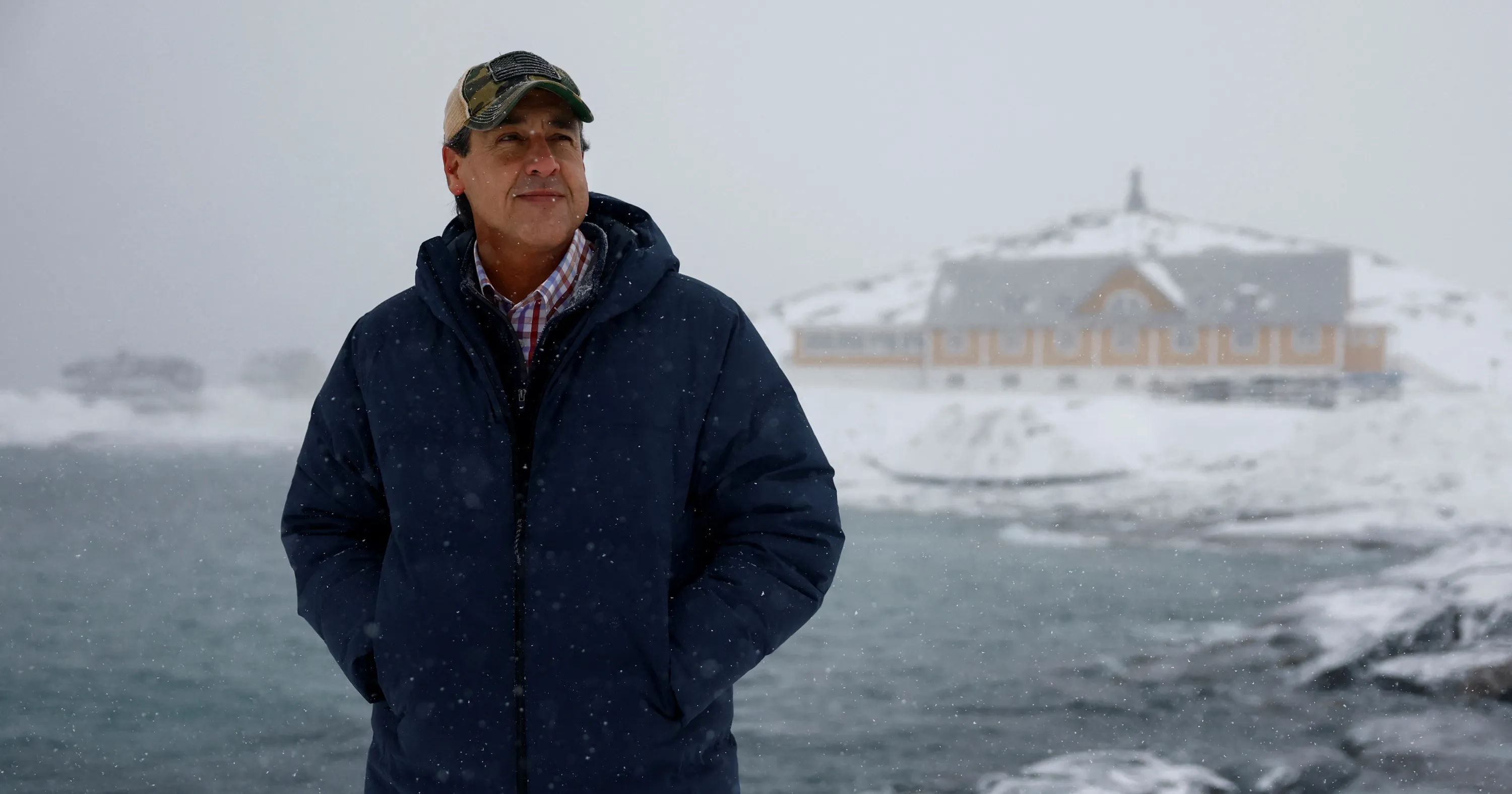 Man in winter coat and cap stands amid falling snow with a large building behind him.