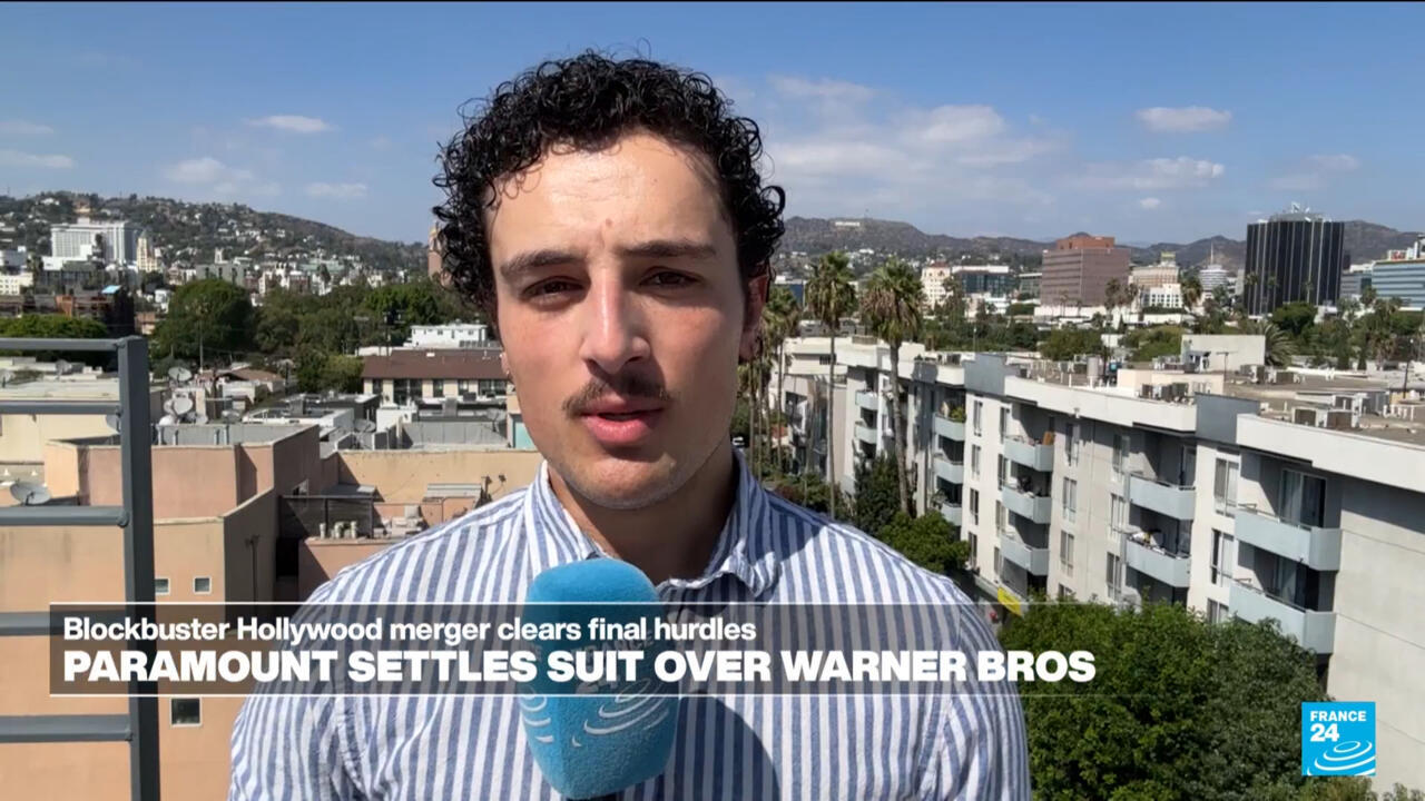 Man holding a blue microphone reports outdoors with a city skyline and hills behind him.