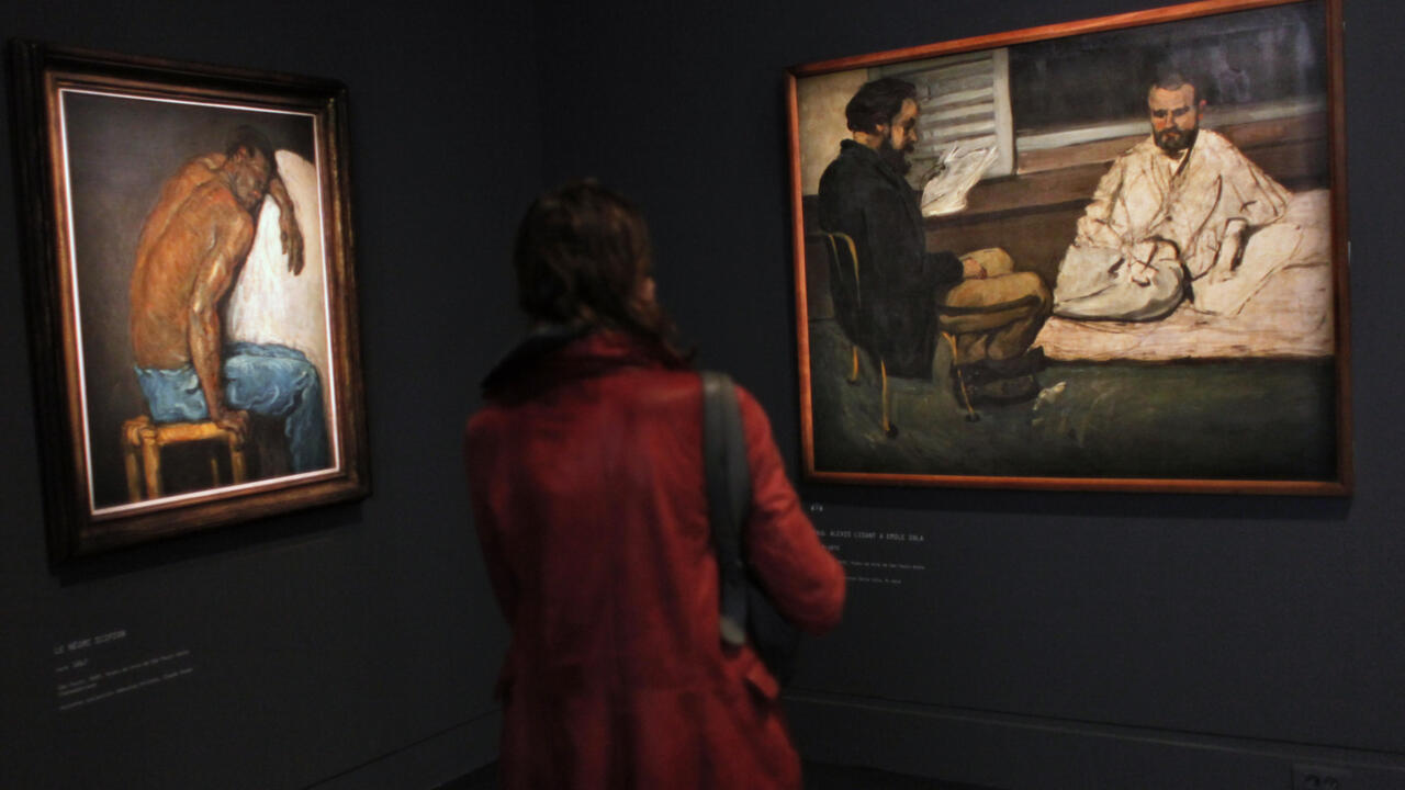 A visitor in a red coat views two framed paintings in a dark gallery.