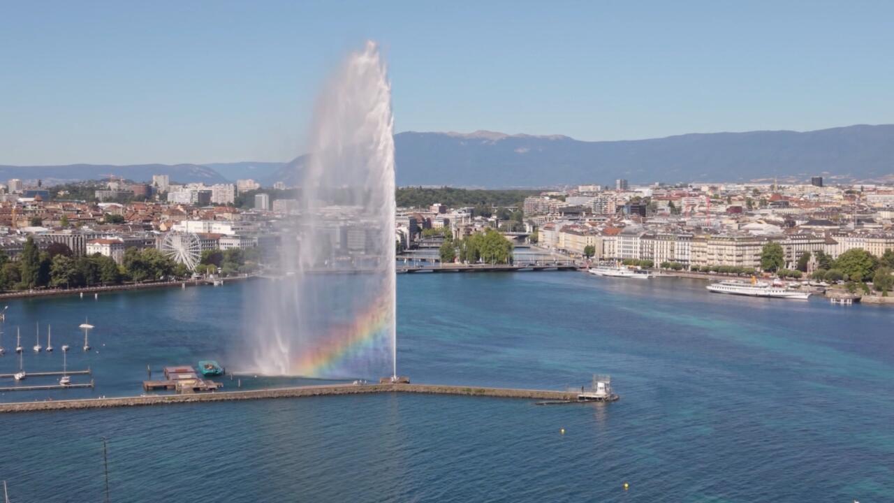 A tall fountain sprays above a lake, with boats, a waterfront city, and distant hills behind it.