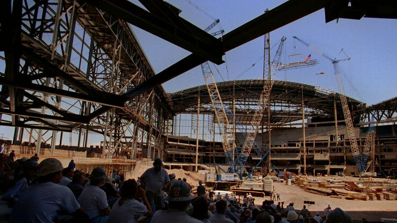 Construction workers watch cranes and steel framing rise at a large stadium construction site.
