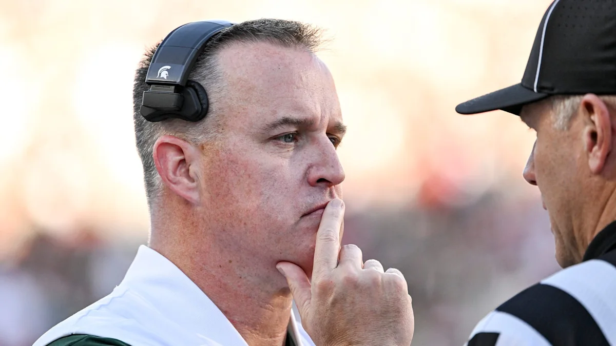 A man wearing a headset touches his lips while facing another man in a cap and striped shirt.