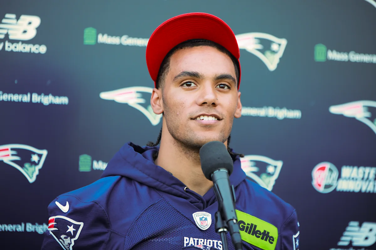 Football player in Patriots jersey and red cap speaking into a microphone at a press conference