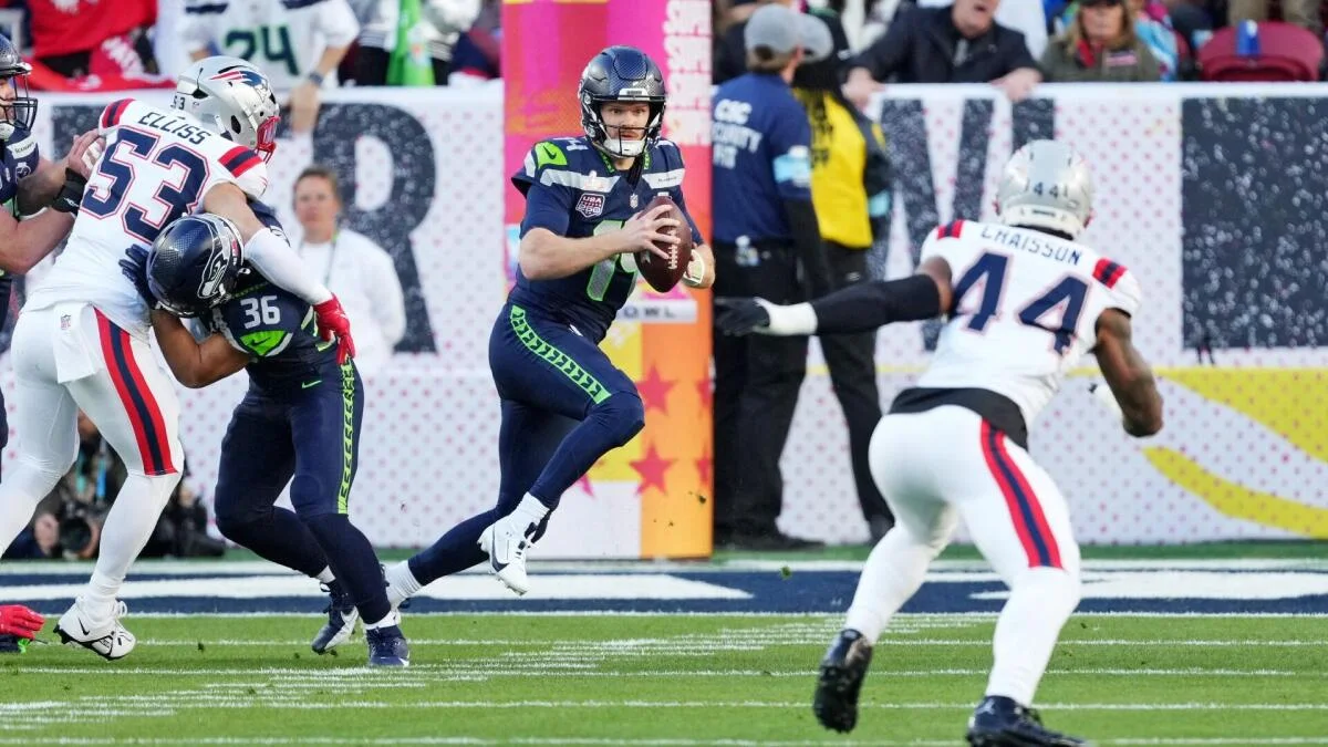 Seattle quarterback throws a pass as Patriots defenders pursue during an NFL game.