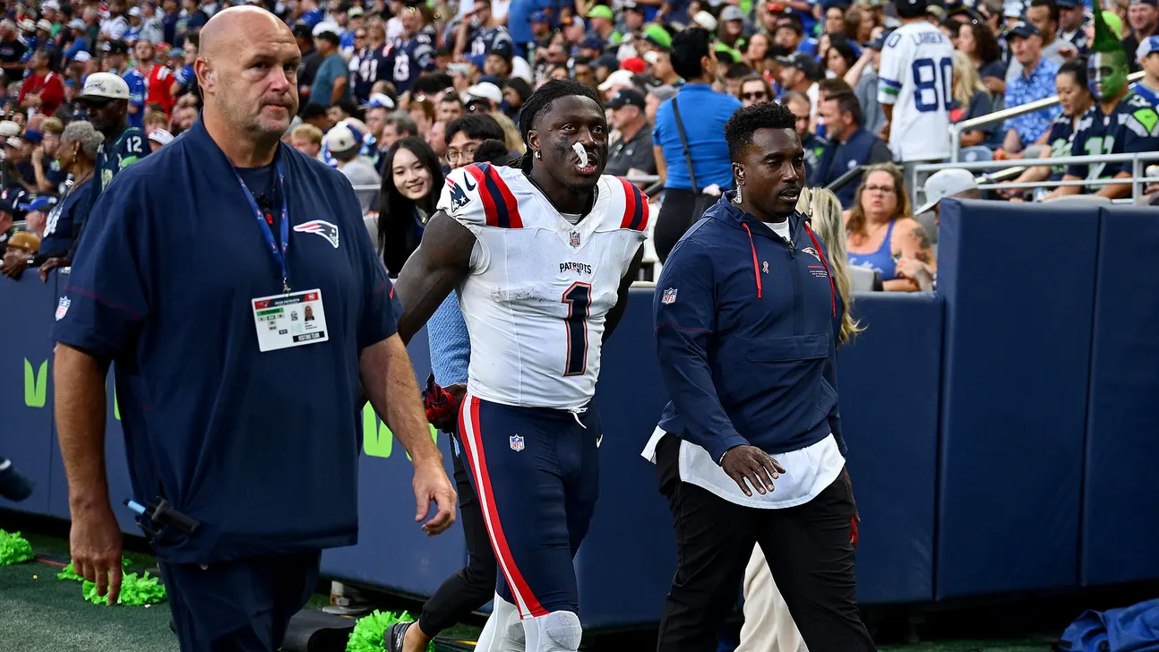 Patriots player in uniform walks off the field with two staff members.
