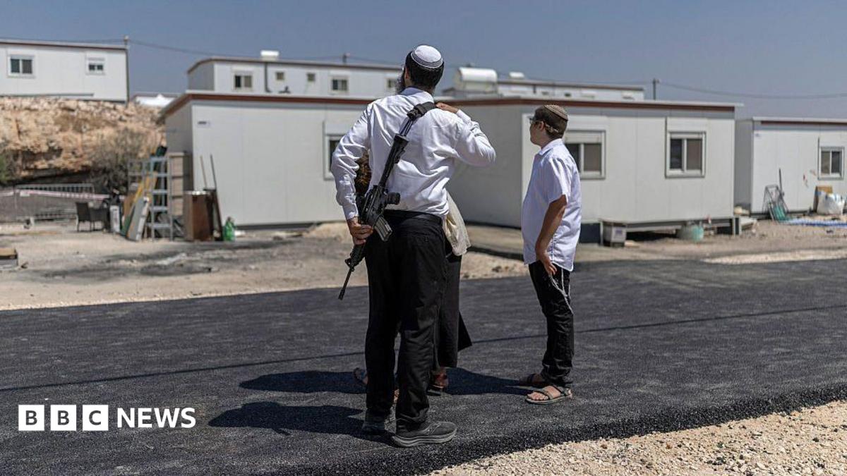 Armed man and boy stand outside temporary buildings on a newly paved road