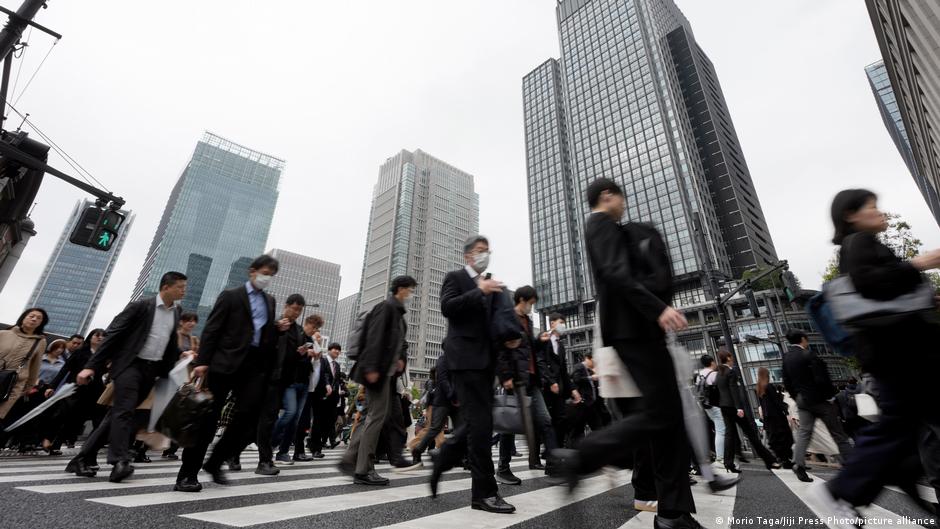 Commuters cross a busy city intersection beneath towering glass office buildings.