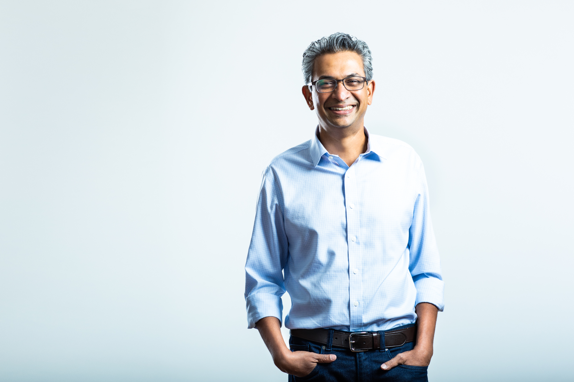 Smiling man with glasses in a light blue shirt stands against a pale background.