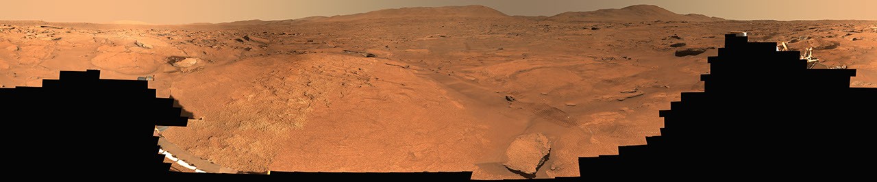 Panoramic view of an orange, rocky Martian landscape beneath a hazy sky, framed by dark foreground silhouettes.