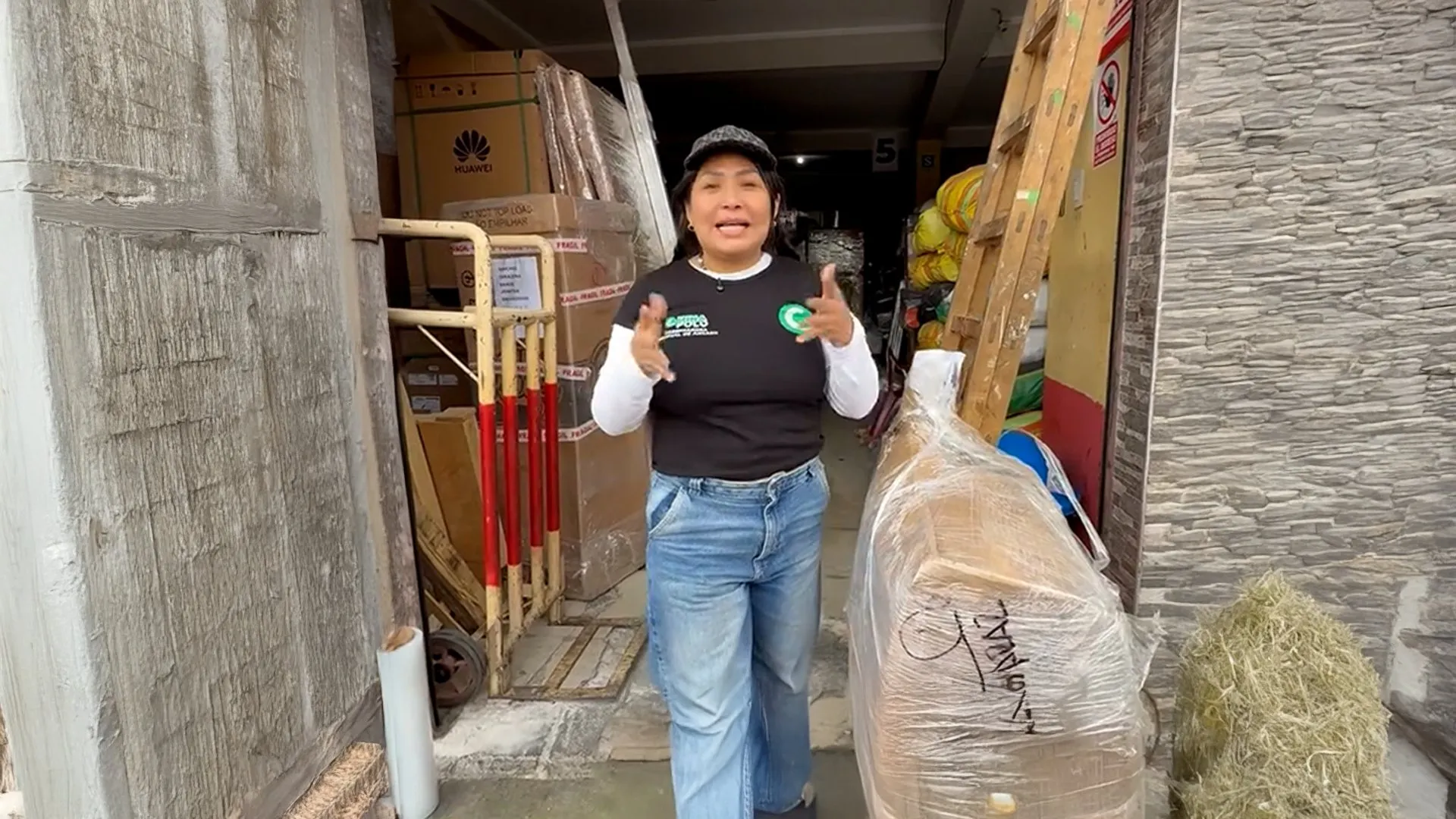 Woman gesturing in a cluttered storage doorway beside boxes, wrapped furniture, and a hand truck