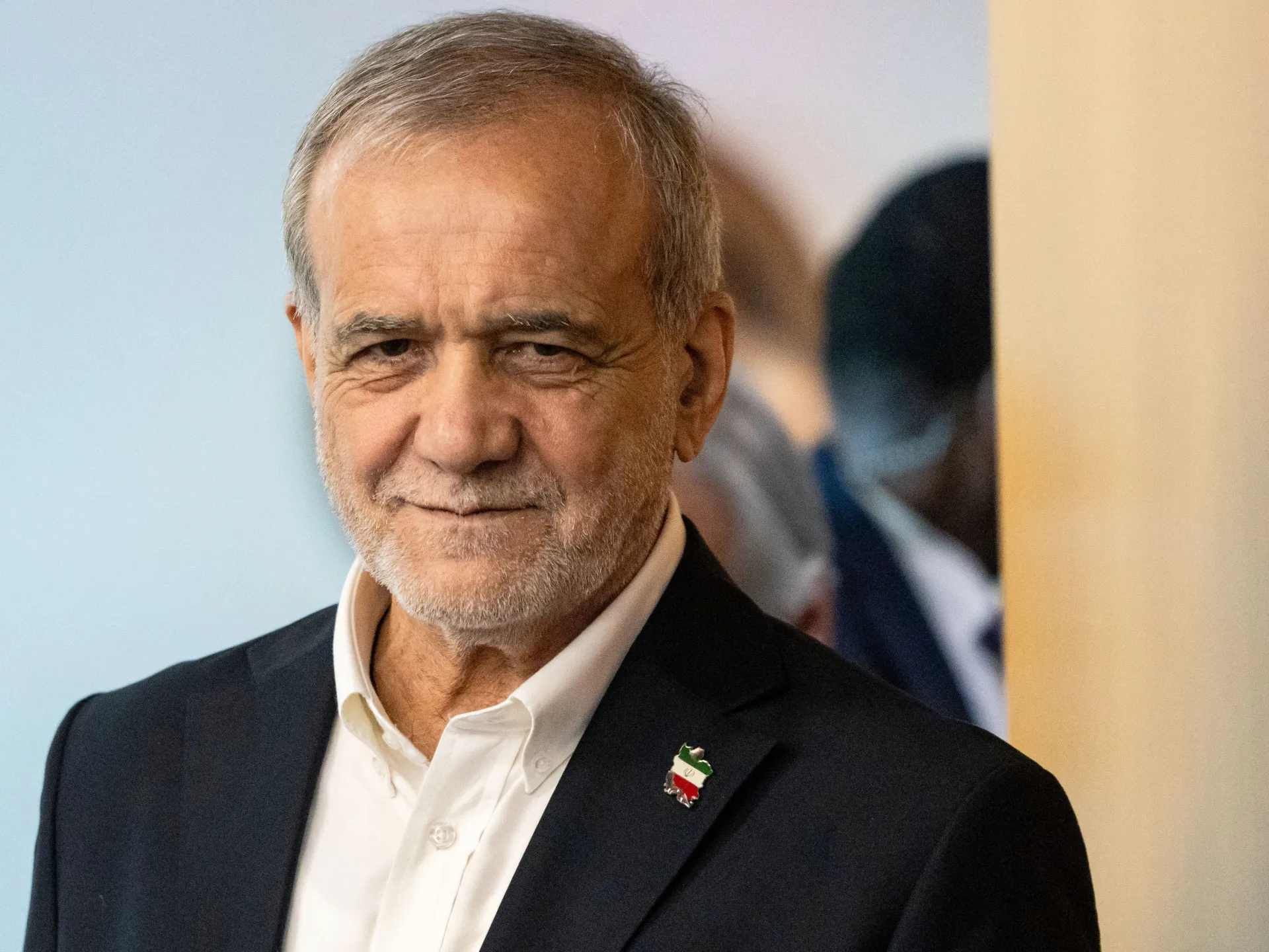 An older gray-haired man in a dark suit and white shirt, wearing an Iran-shaped tricolor lapel pin.