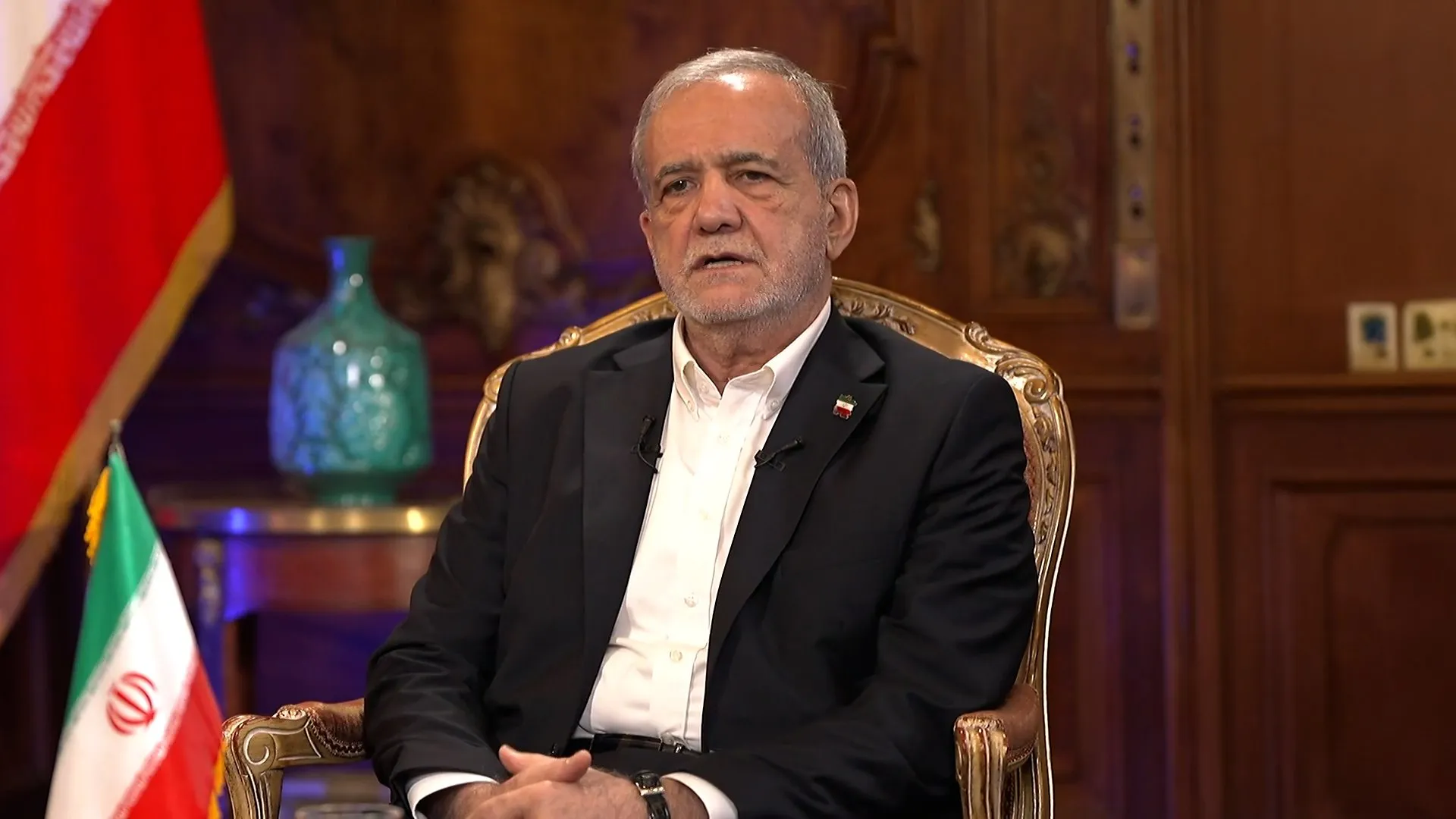 Man in a dark suit sits in an ornate chair beside Iranian flags in a wood-paneled room.