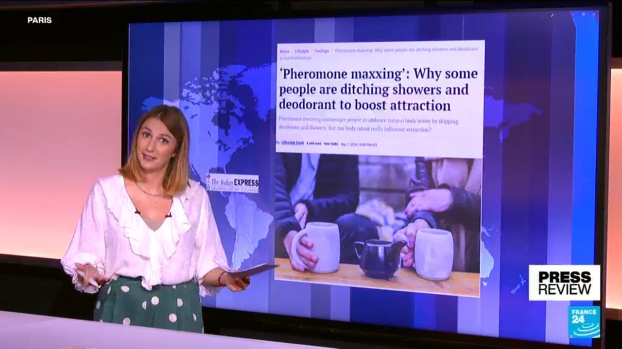 Television presenter stands beside a screen displaying a headline about pheromone maxxing and skipping showers.