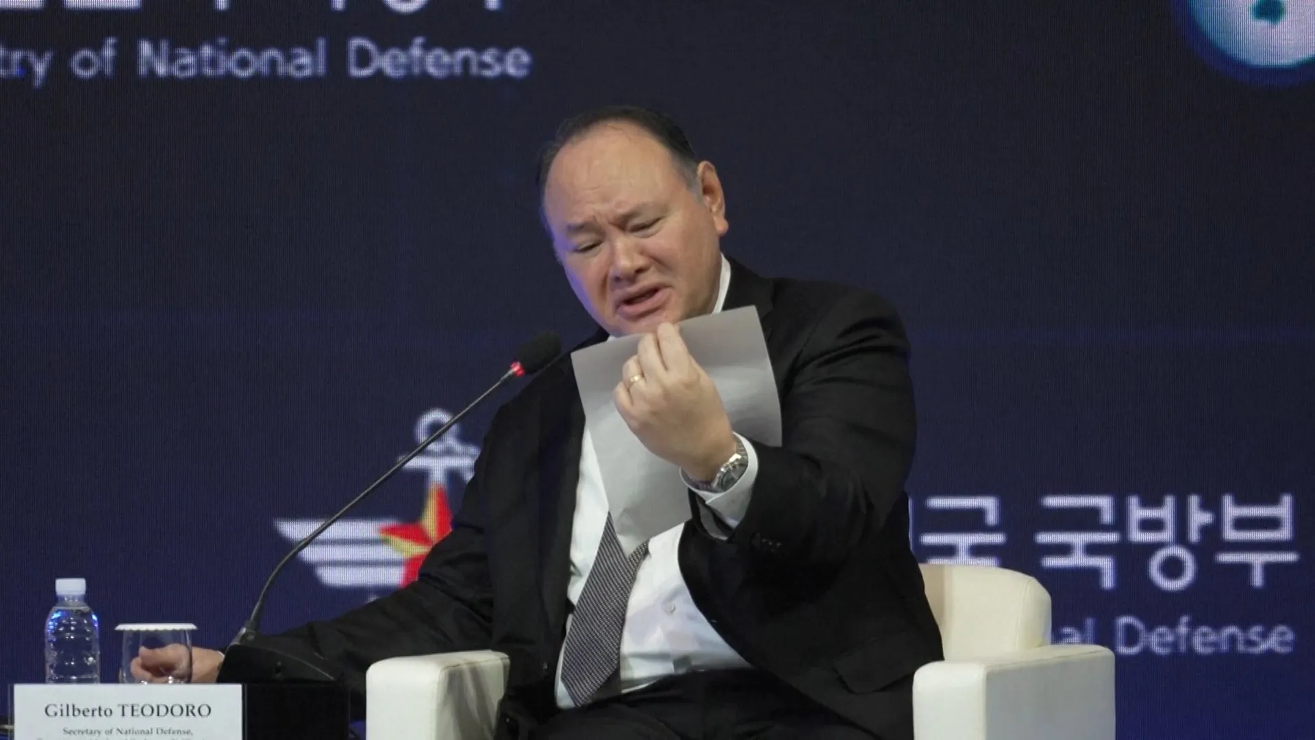 Man in suit holds a sheet of paper while speaking at a panel discussion beside a microphone.