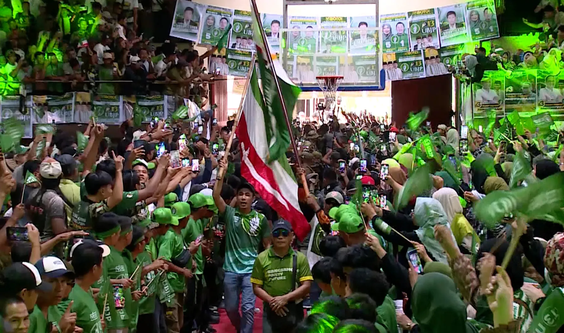 Crowd waves green flags and campaign posters during a packed political rally.