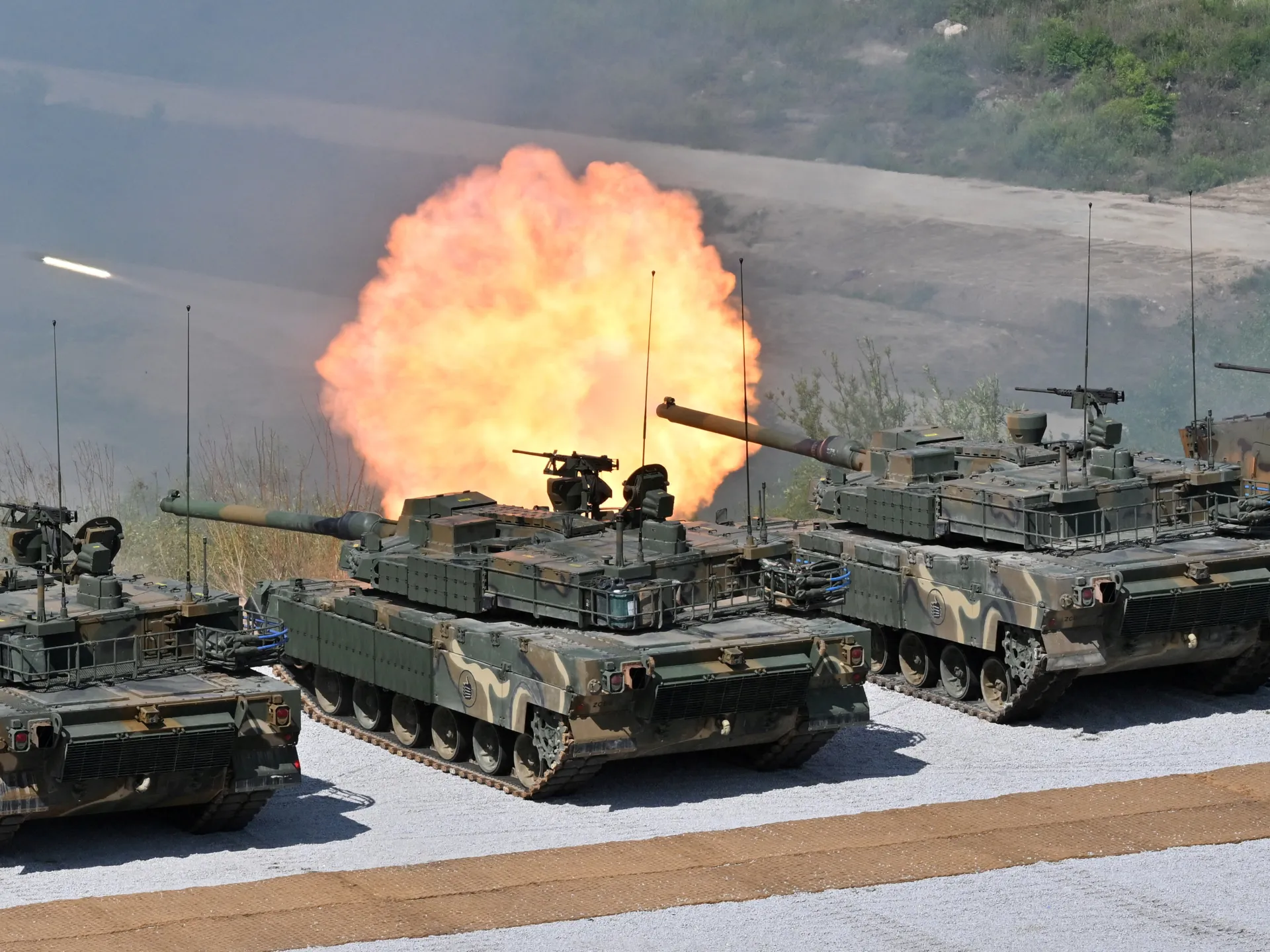 Three camouflaged tanks fire amid a large orange muzzle flash near a hazy waterfront.