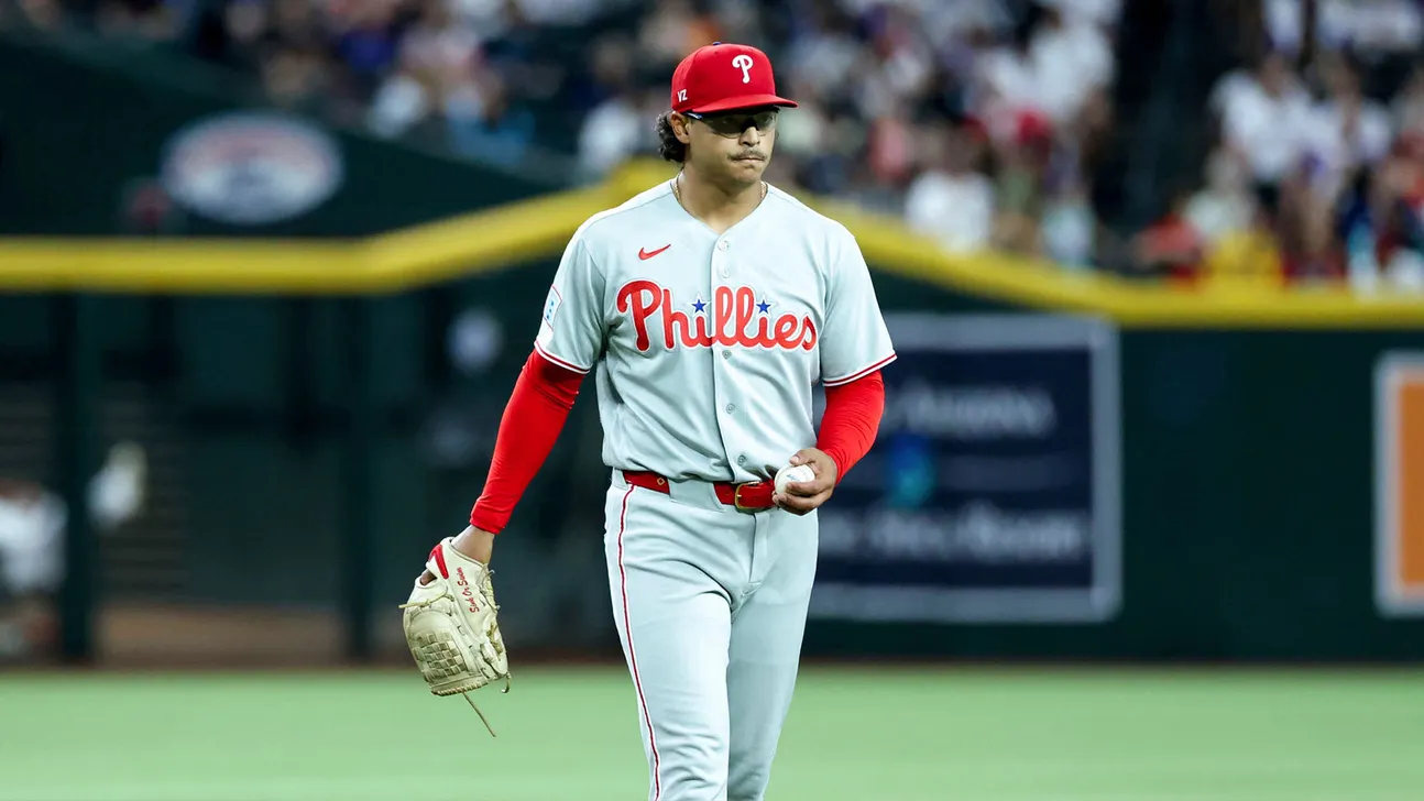 A Phillies pitcher in uniform walks across the field holding a baseball and glove.