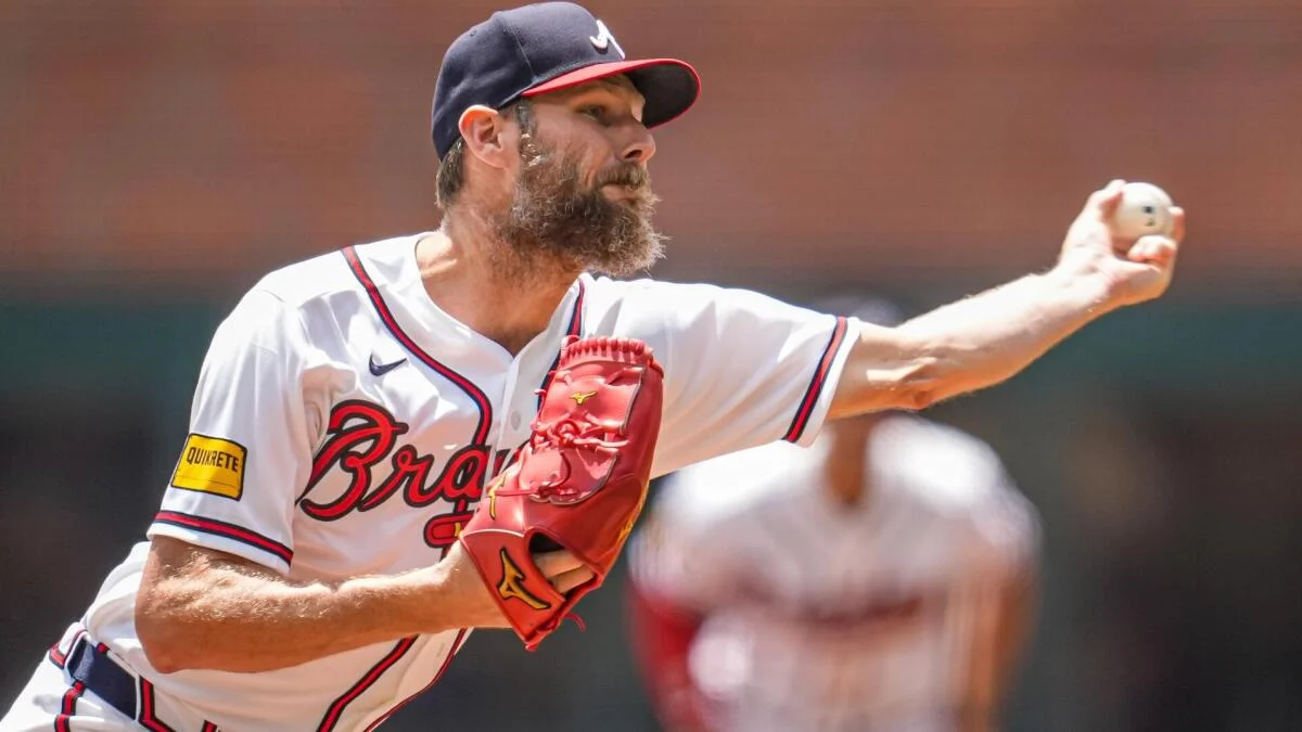 Bearded pitcher in a white Braves uniform extends his arm to throw a baseball.