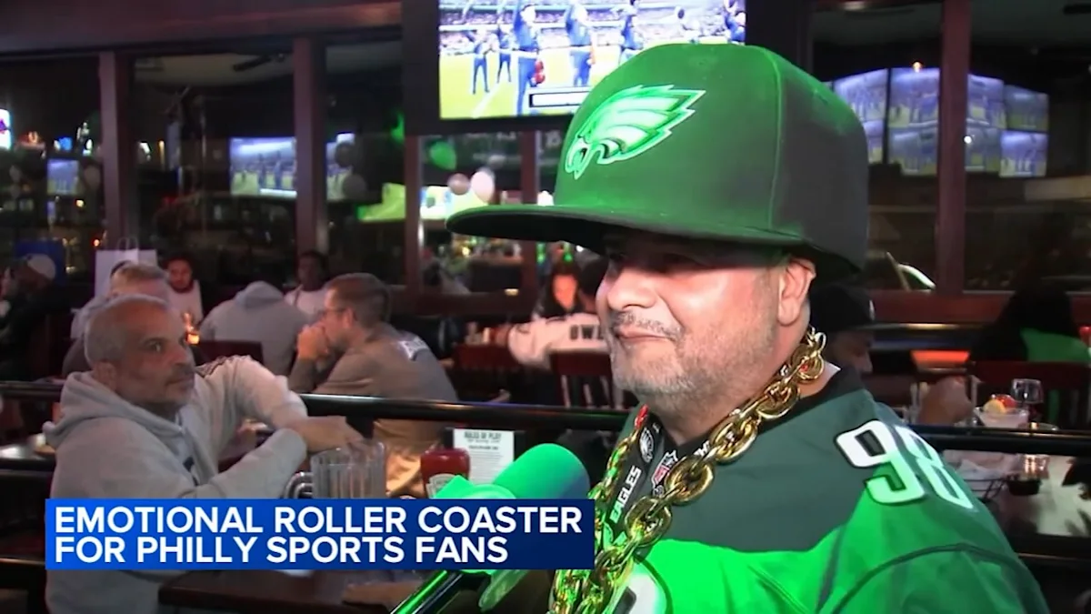 A man in Eagles gear and a large gold chain sits in a crowded sports bar.