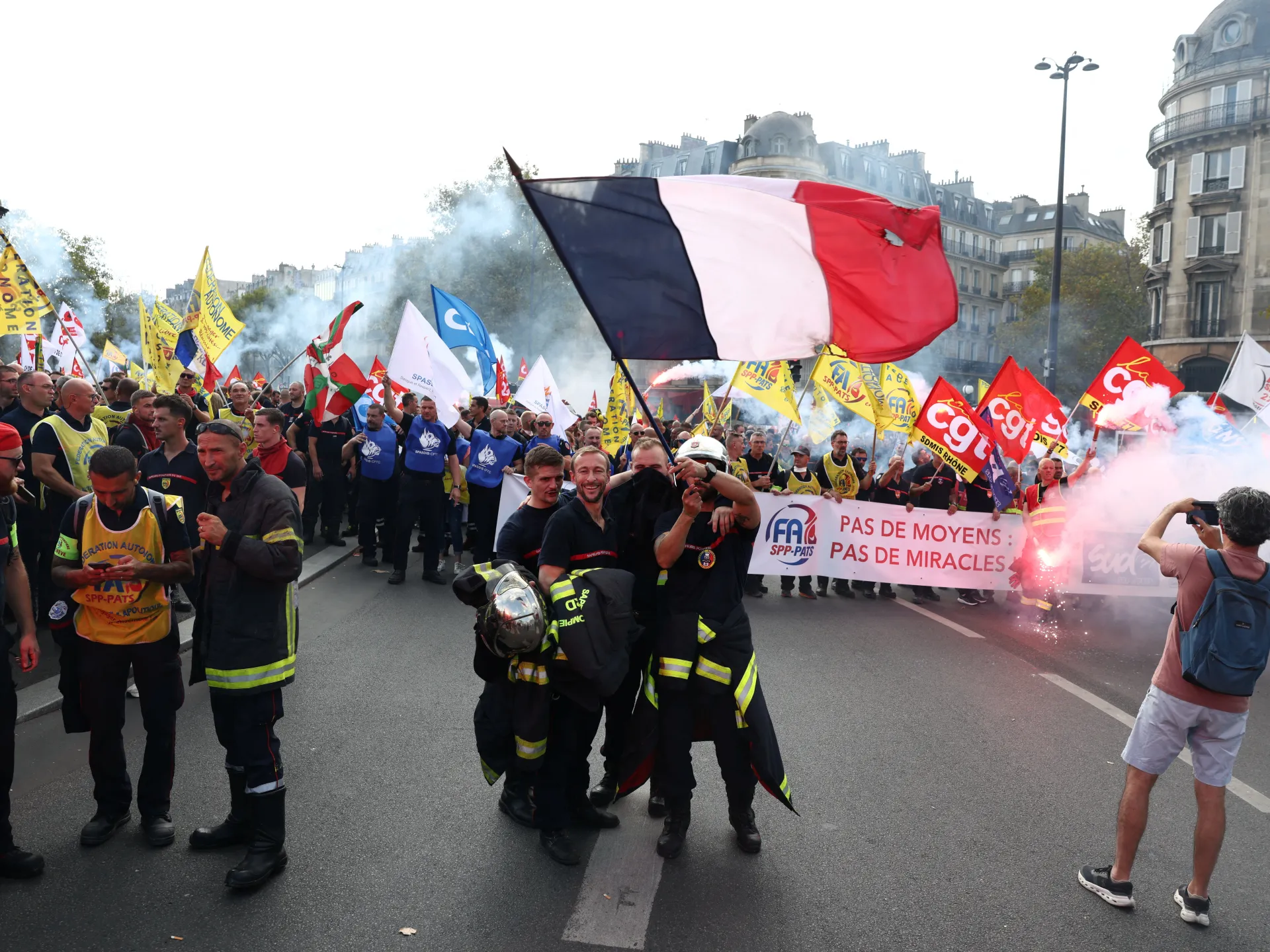 French flag above firefighters and demonstrators carrying union banners amid smoke on a city street.