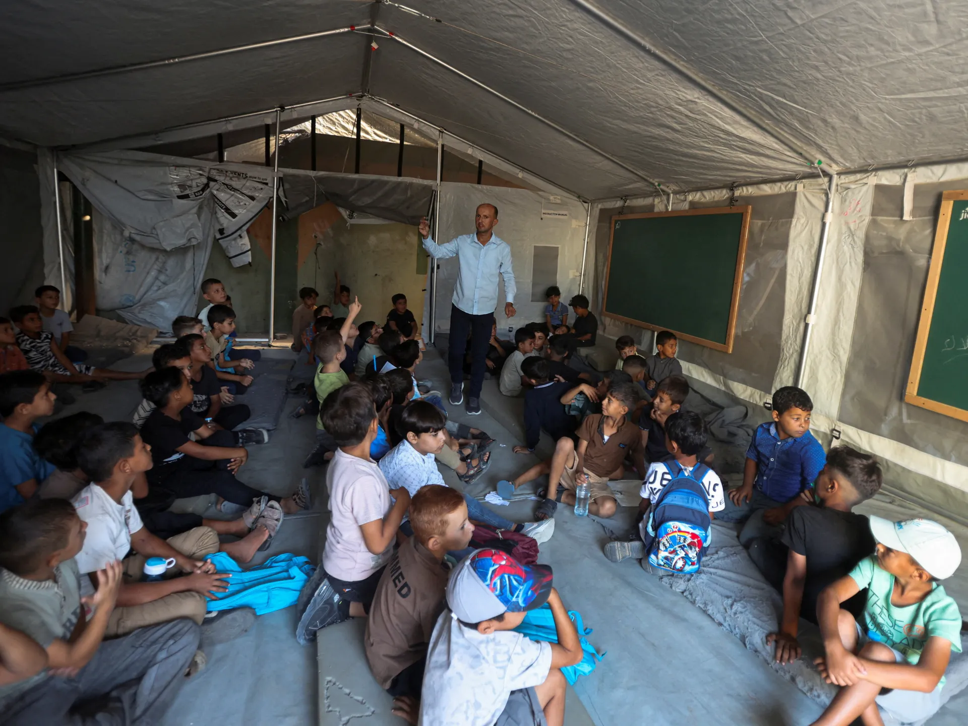 Children sit inside a tent classroom as a teacher stands beside chalkboards.