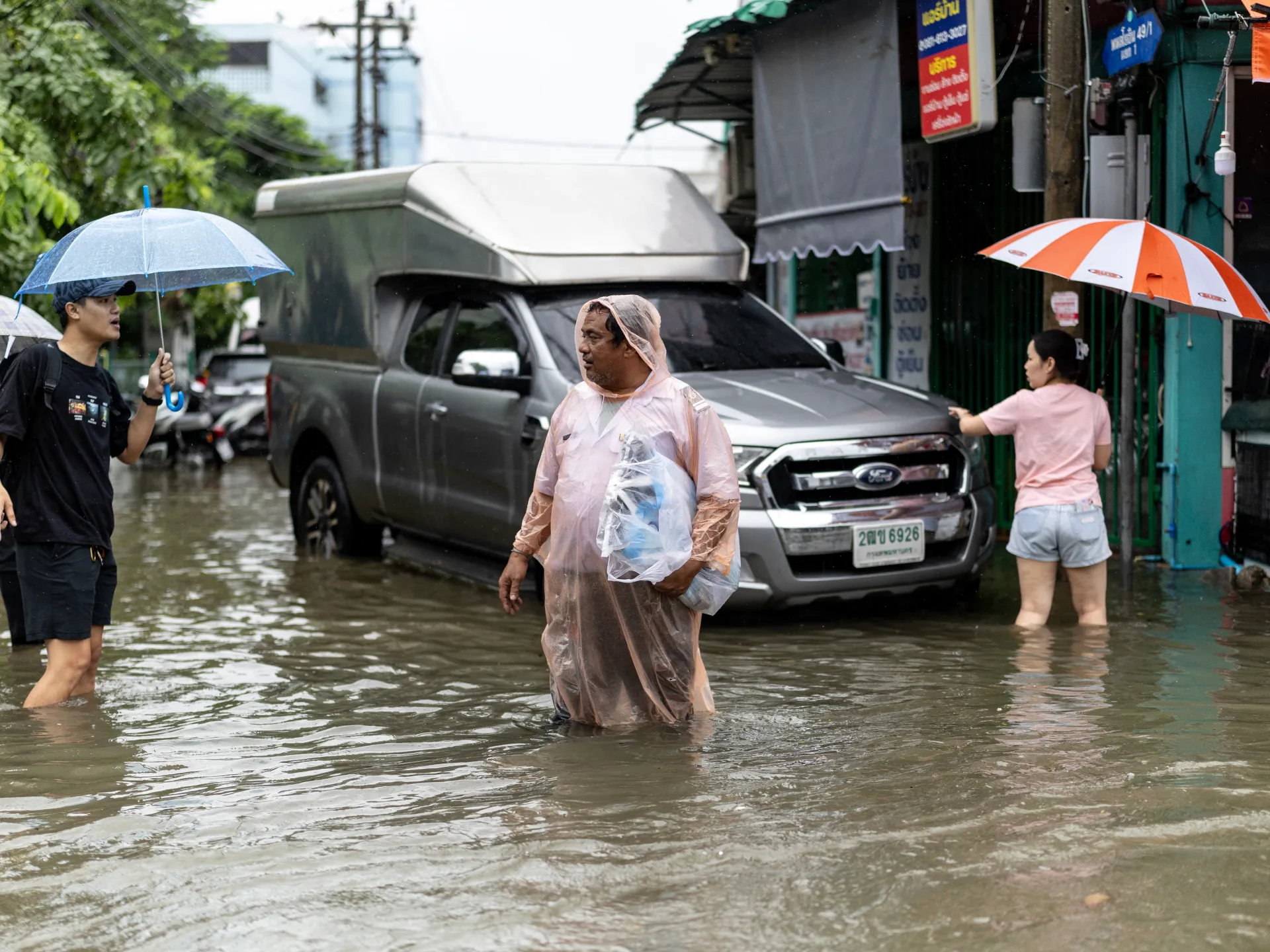 People wade through flooded street water beside a pickup truck, carrying umbrellas in the rain.