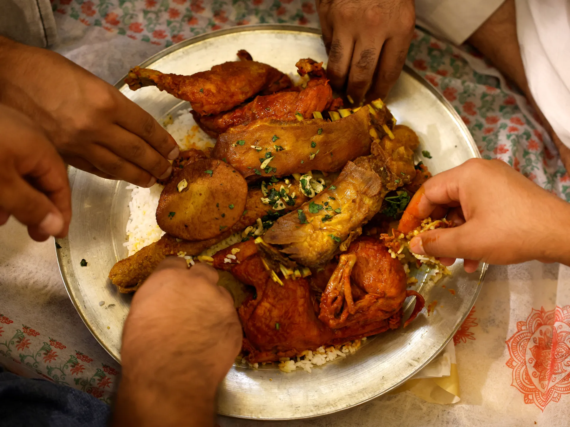 Several hands reach toward a large platter of rice topped with assorted cooked meats and garnishes.