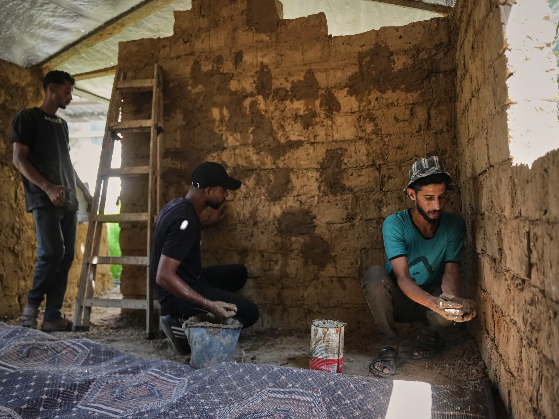 Three men work inside a damaged mud-brick structure, using buckets and hand tools.