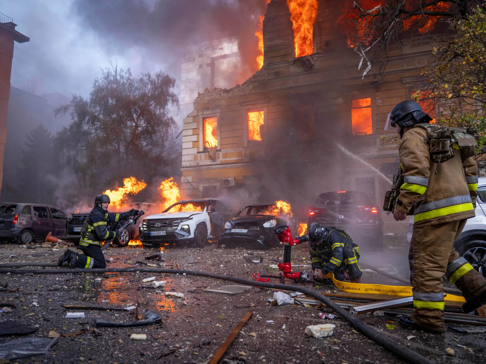 Firefighters battle flames in burning cars and a smoke-filled building as debris covers the street.