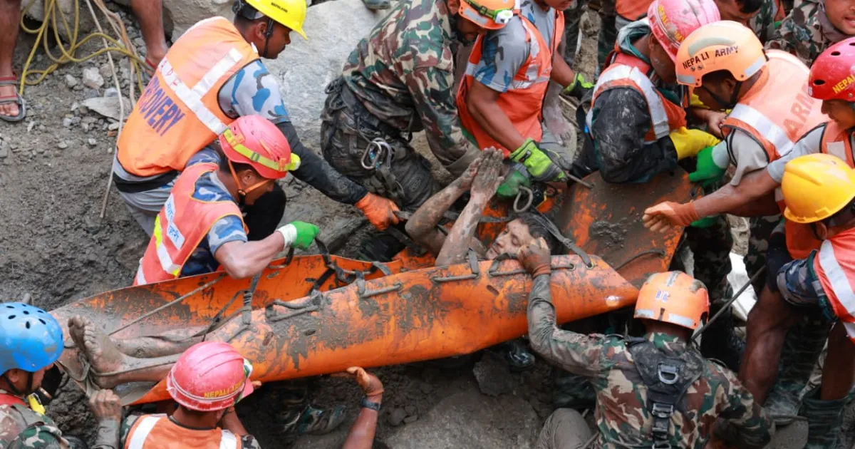 Rescue workers in helmets lift a mud-covered person on an orange stretcher.