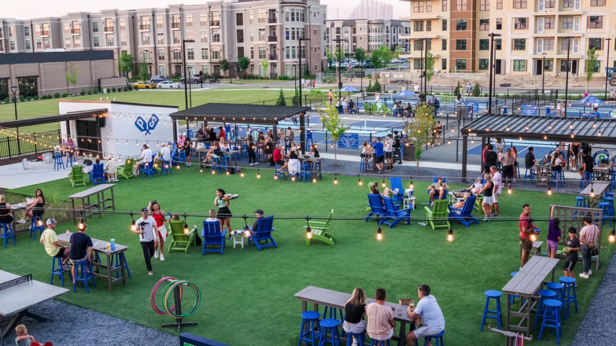 Outdoor pickleball courts and dining area with people socializing among tables, chairs, and string lights.