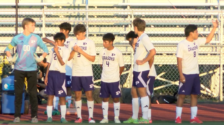 Pittsburg soccer players in white uniforms and a teal-clad goalkeeper stand along the sideline.