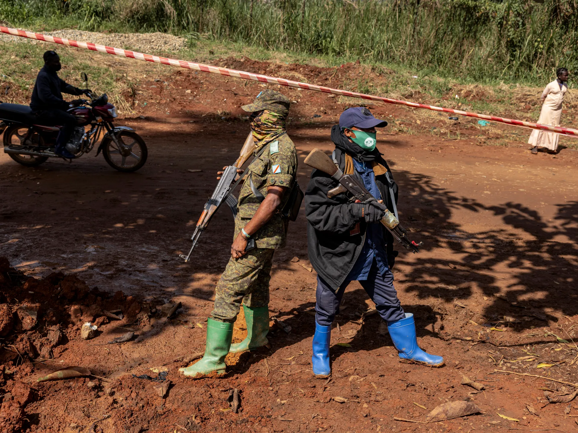 Two armed men in boots stand on muddy ground near a road cordoned with red and white tape.