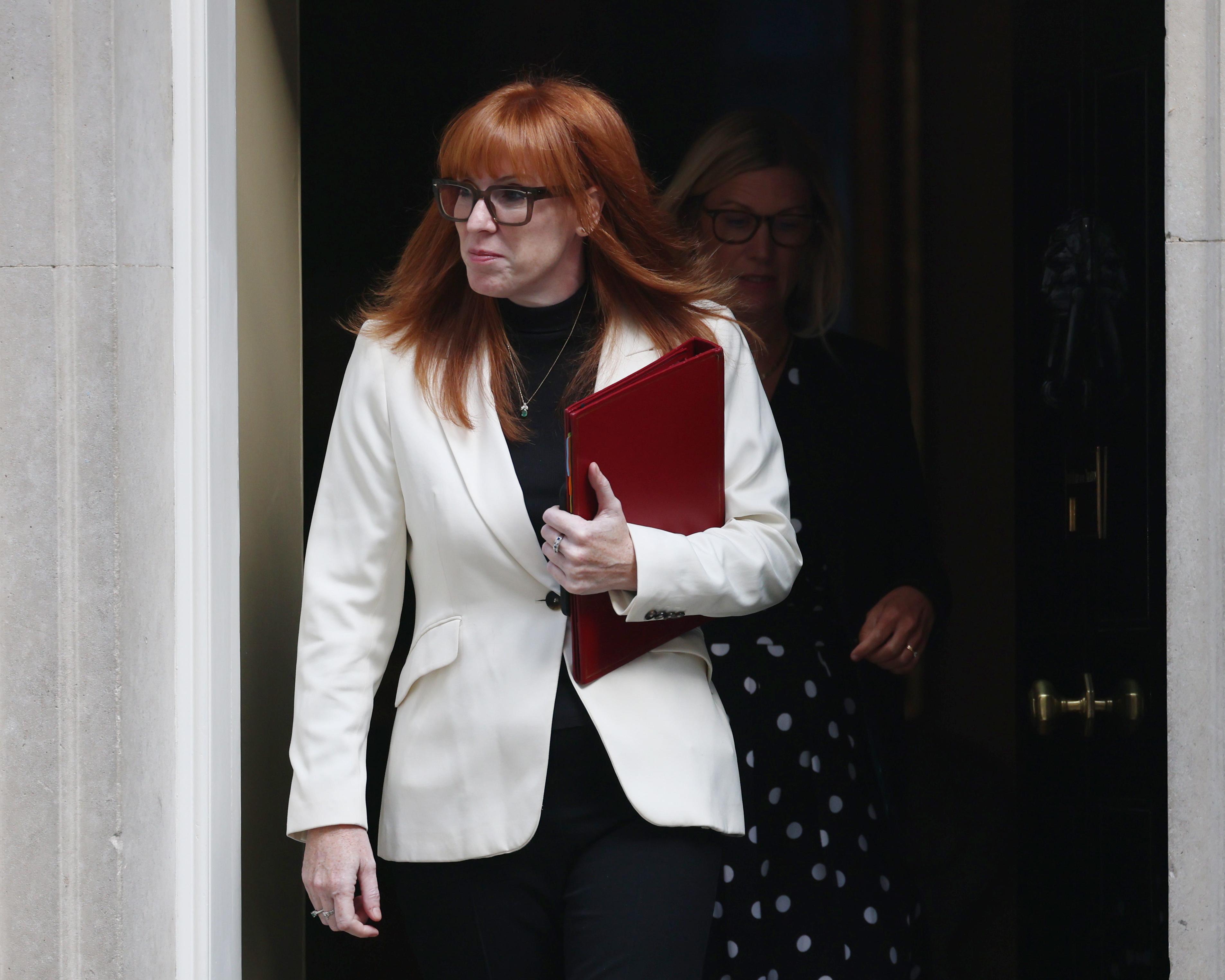 Red-haired woman in glasses exits a doorway holding a red folder, with another woman behind her.