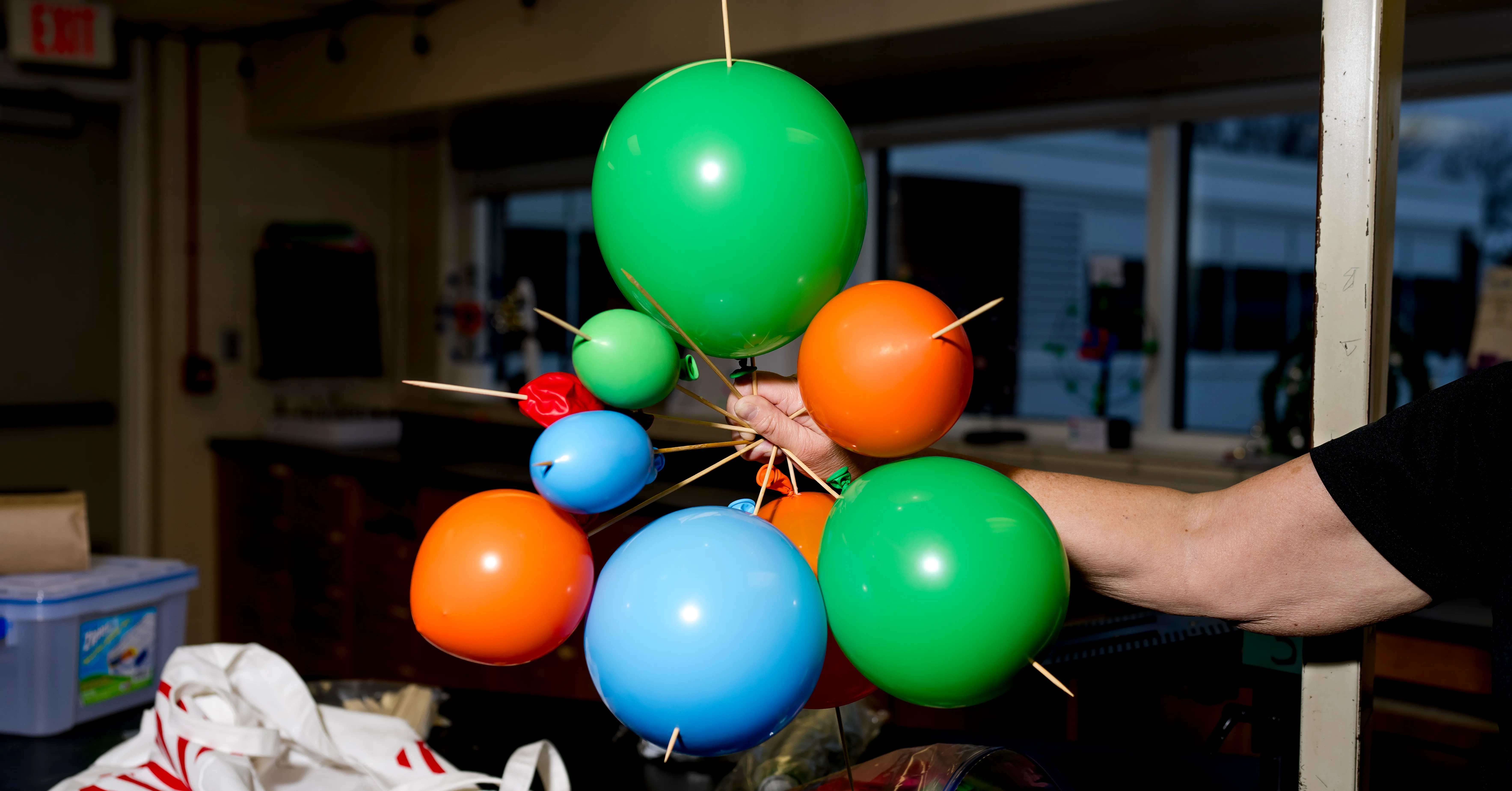 Person holds a colorful cluster of balloons mounted on wooden sticks indoors.
