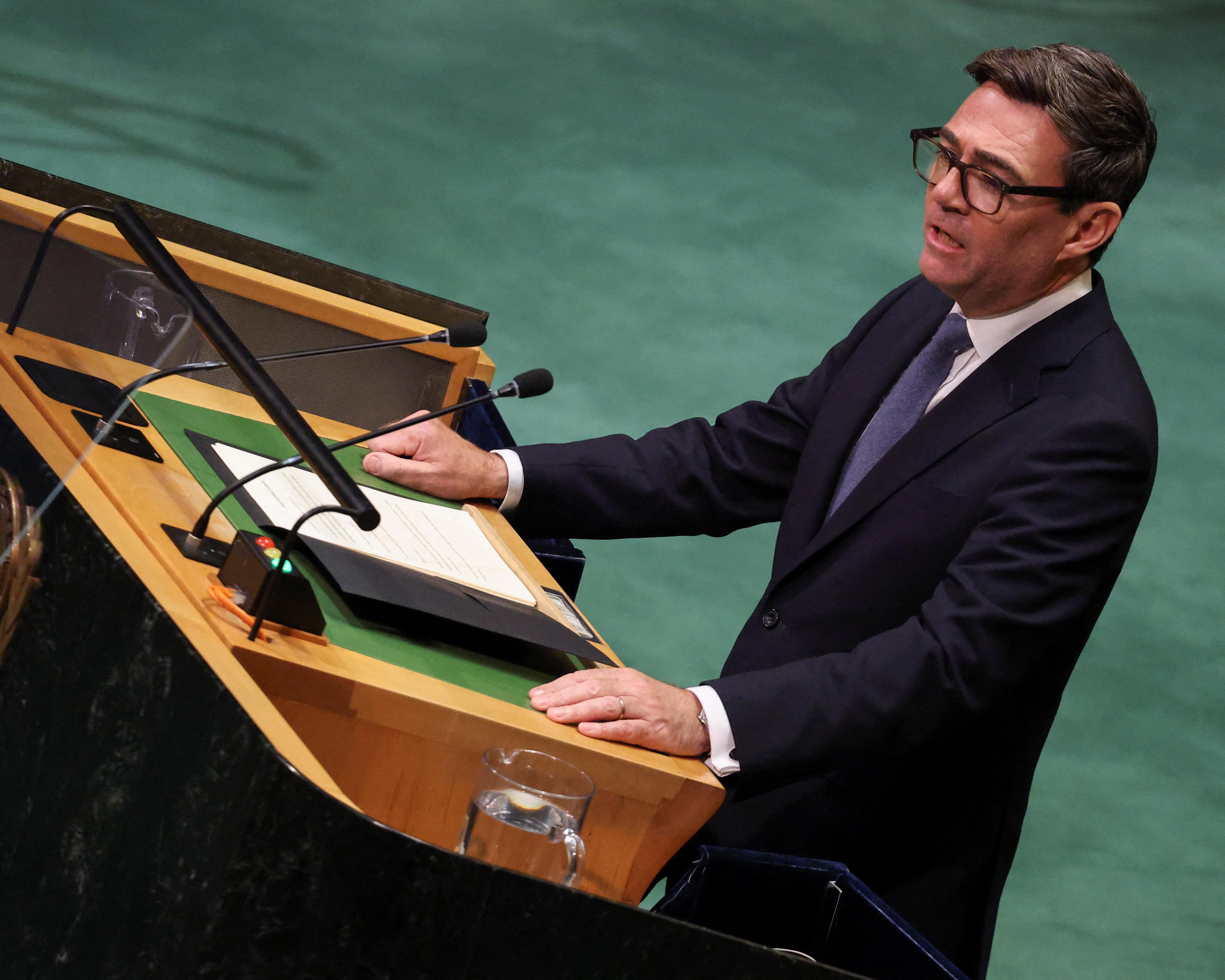 A bespectacled man in a dark suit stands at a wooden lectern with microphones and papers.