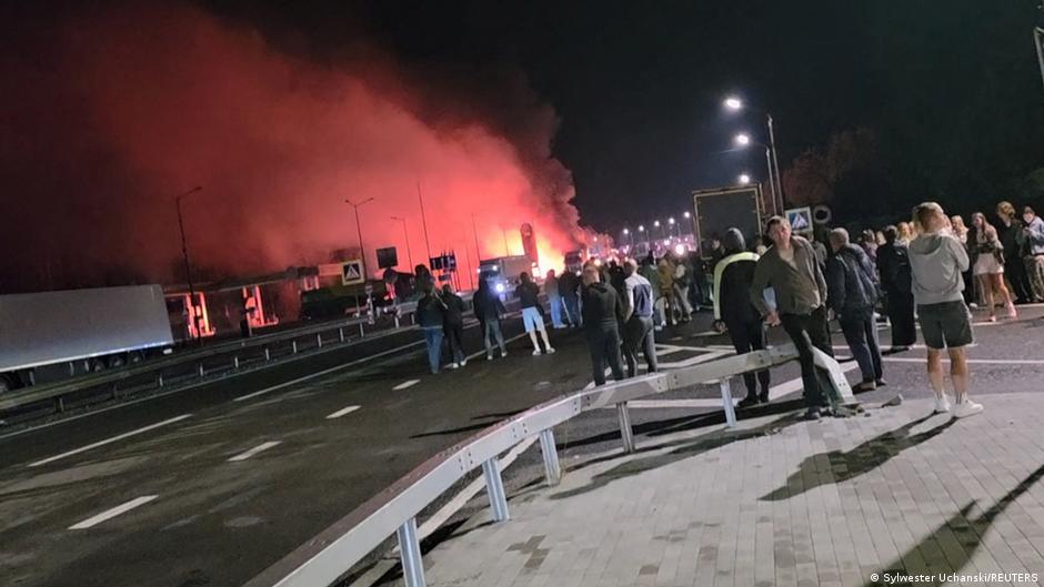 Crowd watches red smoke and flames rising near a roadway at night.
